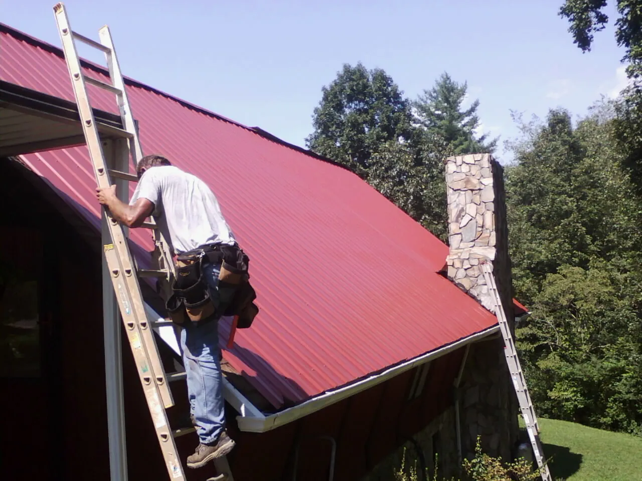 Roofer on a ladder beside a red metal roof and stone chimney — Soffit & Fascia Repair by Roofing Brothers in Norfolk